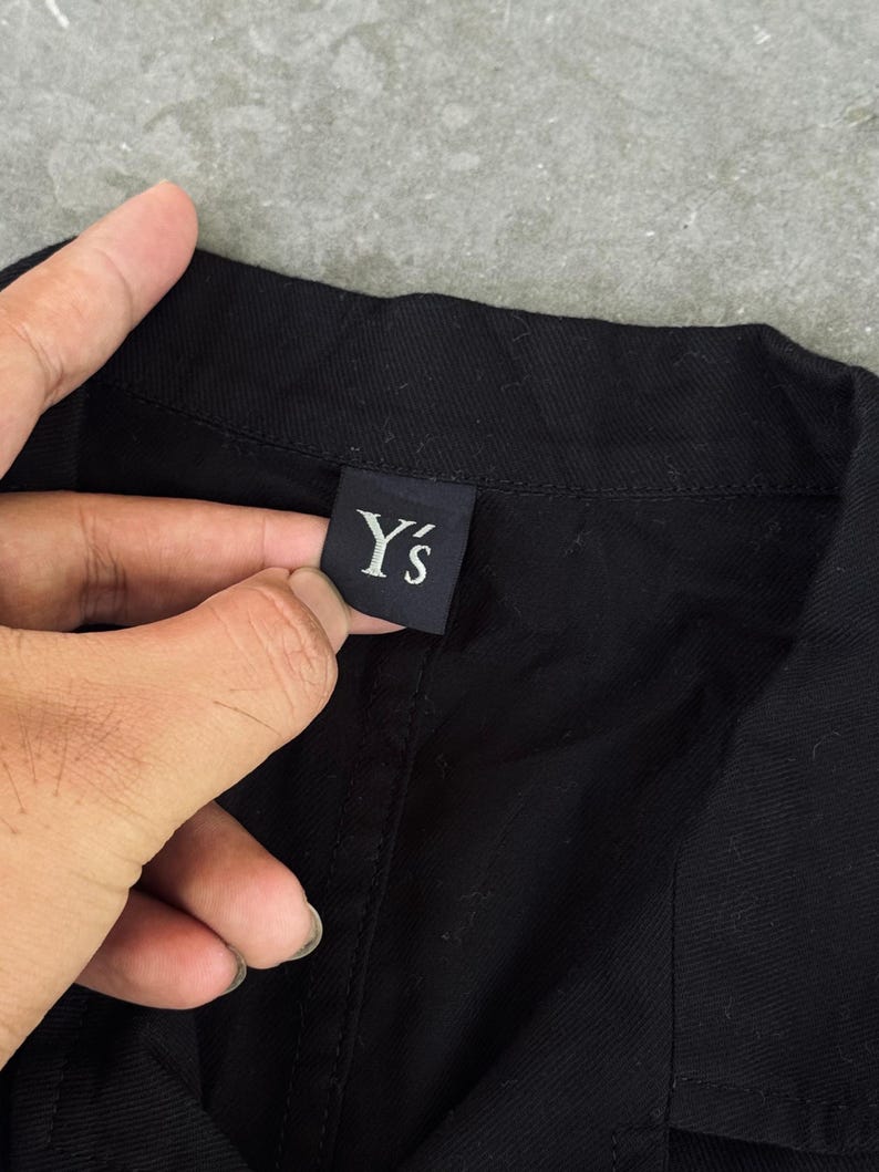 May include: Close-up of a black garment with a label that reads "Y's" in white lettering. The label is rectangular and appears to be made of a dark fabric. The garment is a solid black color.