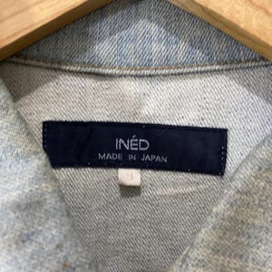 May include: A light blue denim jacket featuring a dark navy blue rectangular label at the inner collar. The label displays the white text INED and MADE IN JAPAN. A small white tag with the number 9 is attached below the main brand label.