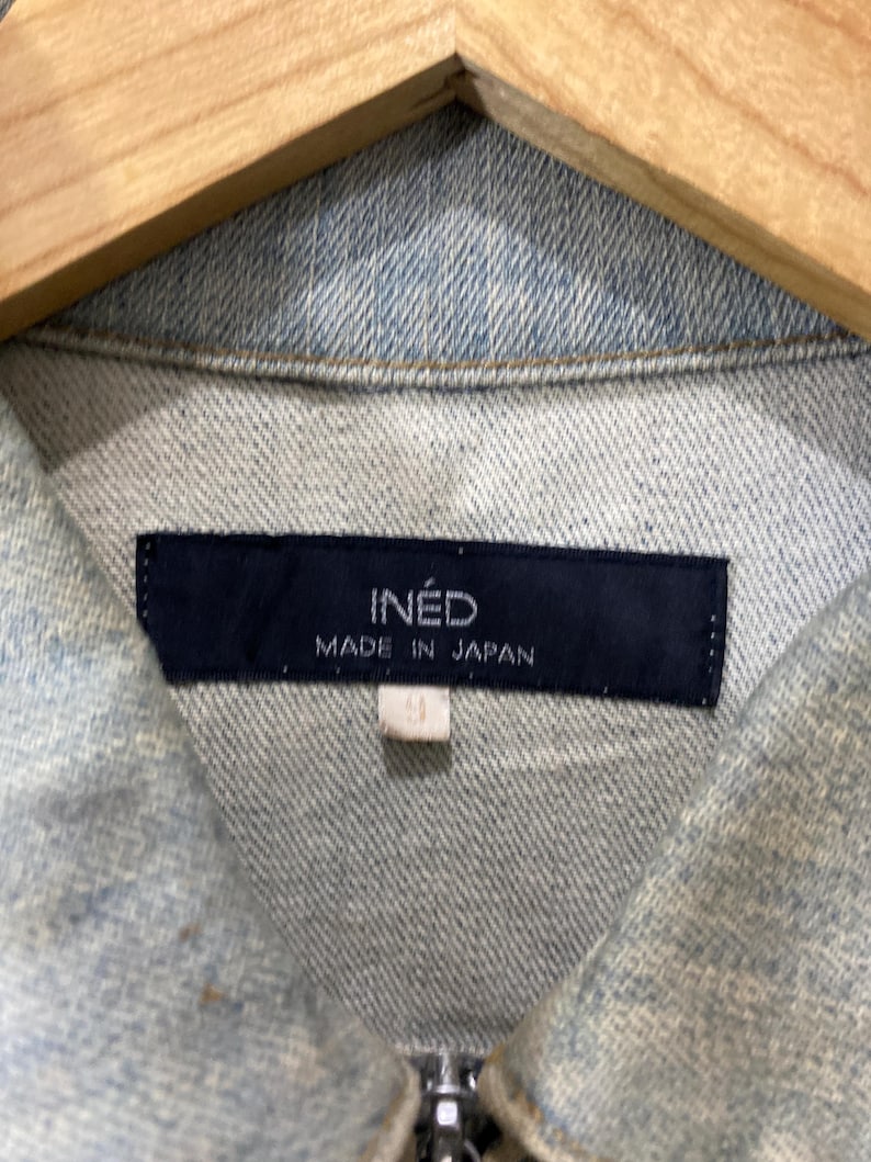 May include: A light blue denim jacket featuring a dark navy blue rectangular label at the inner collar. The label displays the white text INED and MADE IN JAPAN. A small white tag with the number 9 is attached below the main brand label.