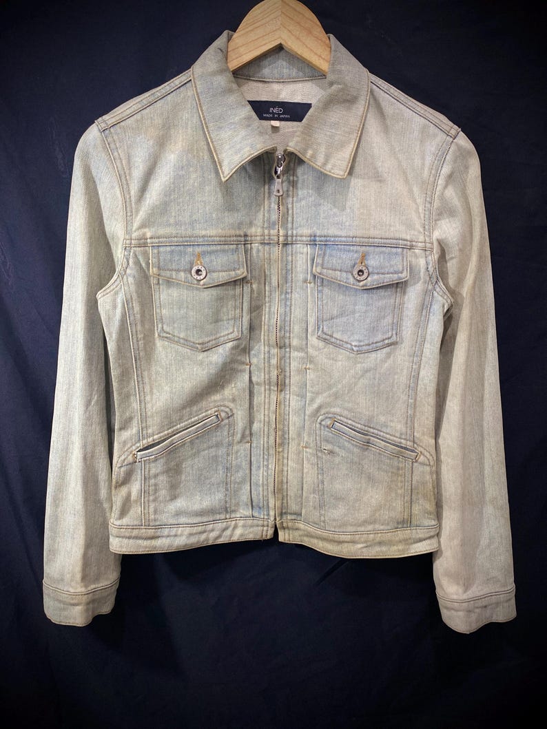 May include: A light-wash blue denim jacket featuring a full-length front zipper, a pointed collar, and long sleeves. The garment includes two chest pockets with buttoned flaps and two lower slanted pockets. The interior label reads INED Made in Japan.