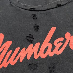 May include: A black t-shirt featuring the word Number printed in a bold red script font. The fabric displays a distressed aesthetic with multiple small, frayed holes scattered across the neckline and the center of the chest area.