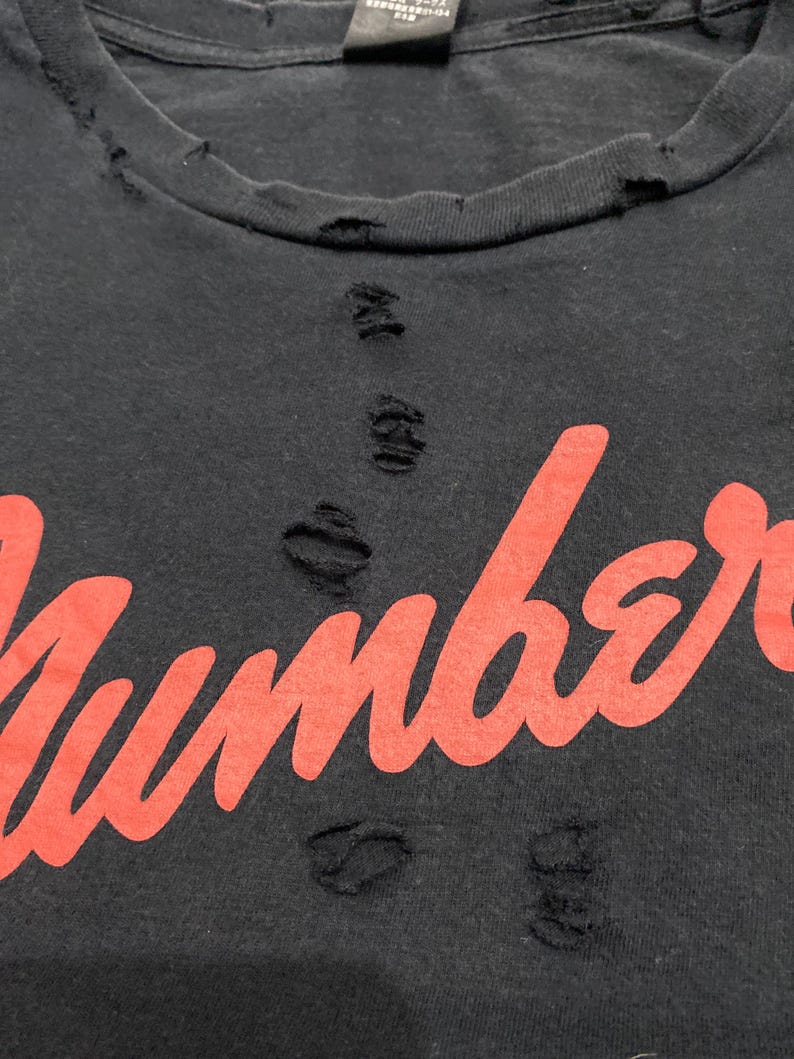 May include: A black t-shirt featuring the word Number printed in a bold red script font. The fabric displays a distressed aesthetic with multiple small, frayed holes scattered across the neckline and the center of the chest area.