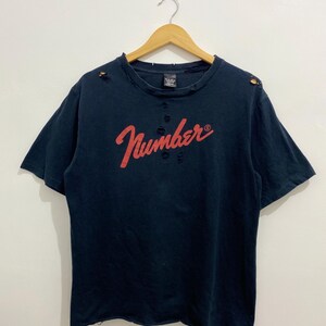 May include: A dark navy blue short-sleeve t-shirt featuring the word Number in red script across the chest. The garment has a distressed finish with small holes around the neckline, shoulders, and hem, and is displayed on a wooden hanger.