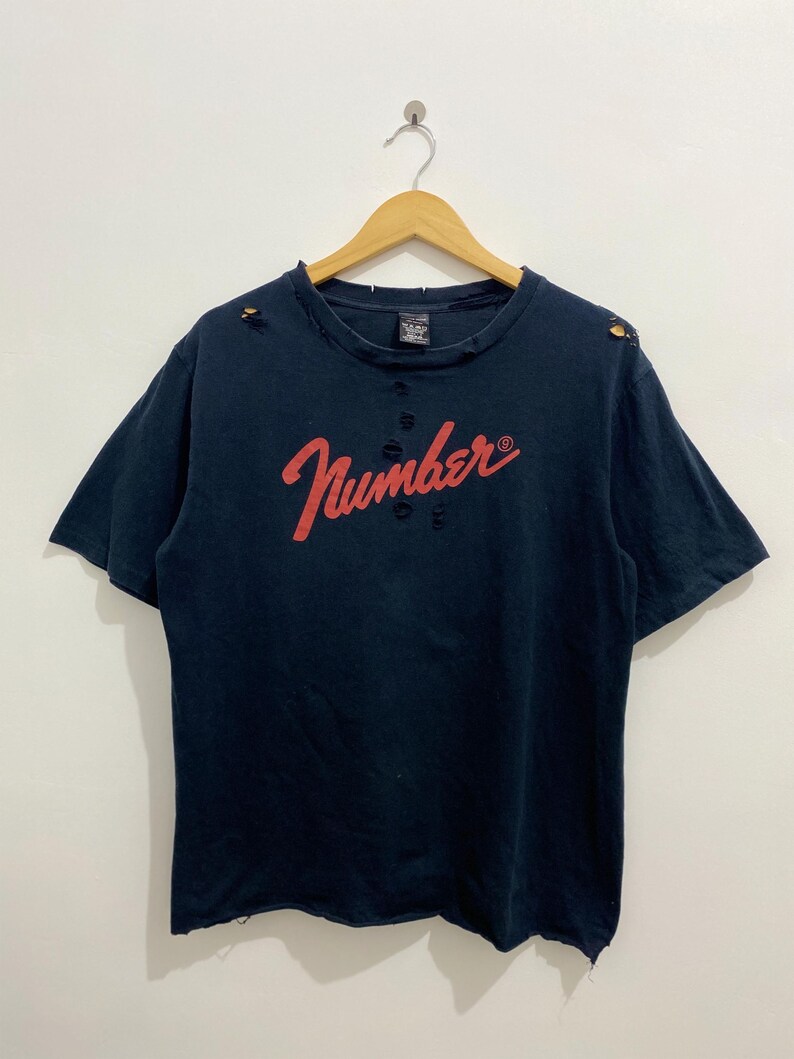 May include: A dark navy blue short-sleeve t-shirt featuring the word Number in red script across the chest. The garment has a distressed finish with small holes around the neckline, shoulders, and hem, and is displayed on a wooden hanger.