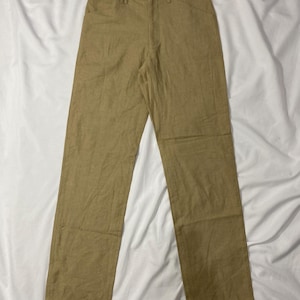 Puede incluir: Pantalones de color beige claro con cierre de bot&oacute;n y trabillas para el cintur&oacute;n. Los pantalones tienen dos bolsillos delanteros y dos traseros. La tela parece ser un material ligero, adecuado para climas c&aacute;lidos. Los pantalones est&aacute;n colocados sobre una superficie blanca.