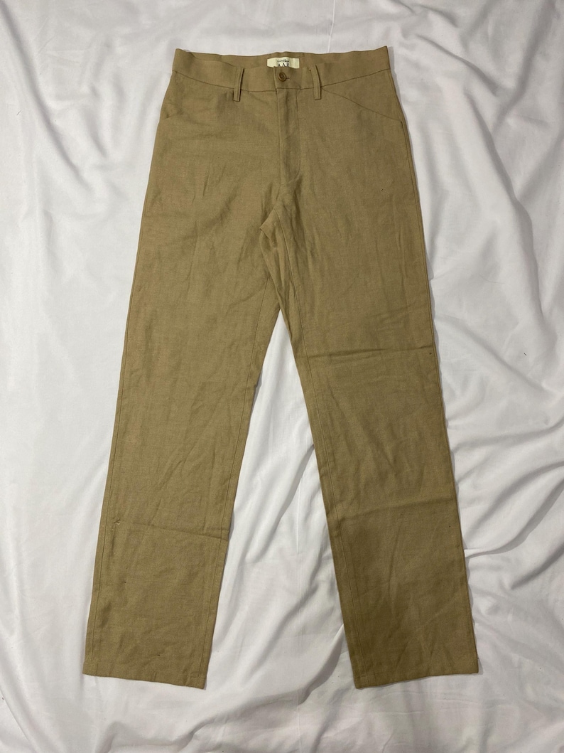 Puede incluir: Pantalones de color beige claro con cierre de bot&oacute;n y trabillas para el cintur&oacute;n. Los pantalones tienen dos bolsillos delanteros y dos traseros. La tela parece ser un material ligero, adecuado para climas c&aacute;lidos. Los pantalones est&aacute;n colocados sobre una superficie blanca.