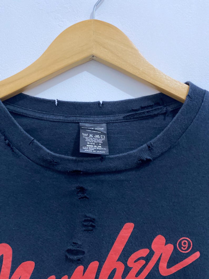 May include: A black t-shirt with a distressed, frayed neckline and small holes throughout the fabric. The shirt features a red graphic print and a black interior tag that reads NUMBER (N)INE 100% COTTON, size 4, and MADE IN JAPAN.
