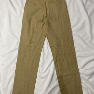 Puede incluir: Pantalones de color beige con un dise&ntilde;o de pierna recta. Los pantalones tienen dos bolsillos traseros y cierre de bot&oacute;n. La tela parece ser un material ligero, adecuado para climas c&aacute;lidos. Los pantalones est&aacute;n colocados sobre una superficie blanca.