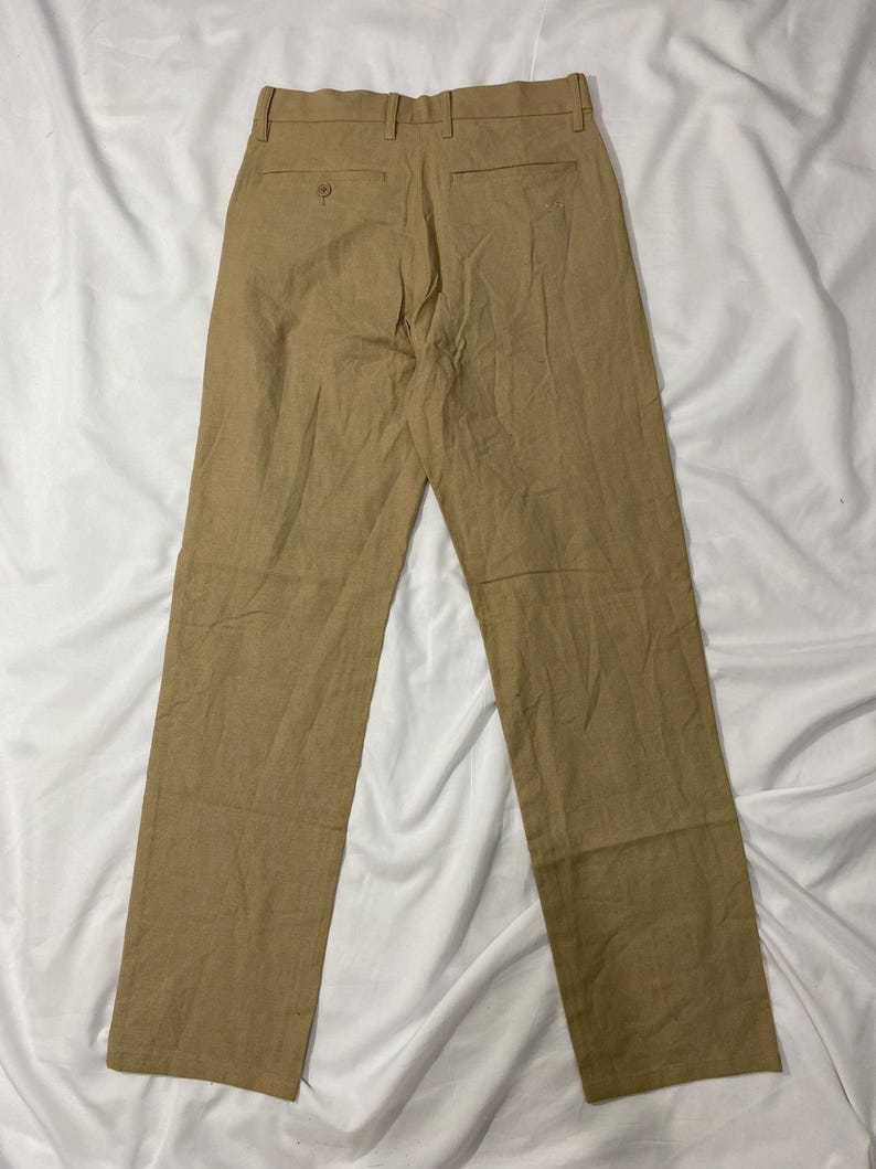 Puede incluir: Pantalones de color beige con un dise&ntilde;o de pierna recta. Los pantalones tienen dos bolsillos traseros y cierre de bot&oacute;n. La tela parece ser un material ligero, adecuado para climas c&aacute;lidos. Los pantalones est&aacute;n colocados sobre una superficie blanca.