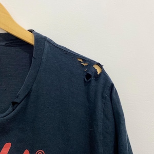 May include: A dark navy blue t-shirt featuring a red graphic print on the chest. The fabric shows visible signs of wear, including small holes and fraying along the shoulder seam and the neckline area, indicating a distressed, aged appearance.