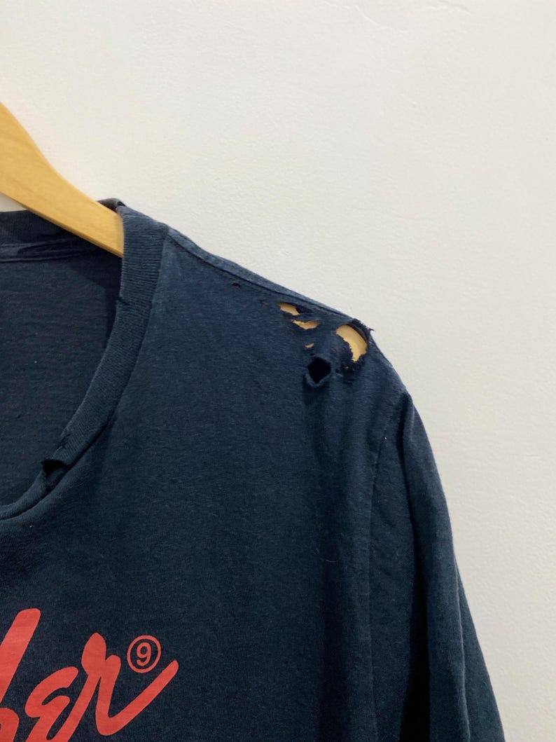 May include: A dark navy blue t-shirt featuring a red graphic print on the chest. The fabric shows visible signs of wear, including small holes and fraying along the shoulder seam and the neckline area, indicating a distressed, aged appearance.