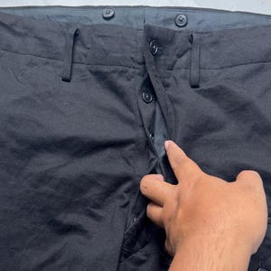 Puede incluir: Primer plano de pantalones negros con cierre de bot&oacute;n y trabillas para el cintur&oacute;n. Los pantalones tienen un dise&ntilde;o cl&aacute;sico con bragueta de botones y un ajuste a medida. La tela parece ser un material de peso medio, adecuado para diversas ocasiones.