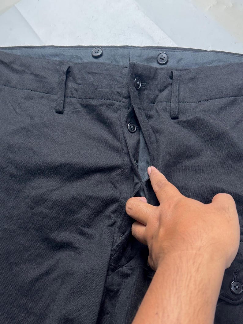 Puede incluir: Primer plano de pantalones negros con cierre de bot&oacute;n y trabillas para el cintur&oacute;n. Los pantalones tienen un dise&ntilde;o cl&aacute;sico con bragueta de botones y un ajuste a medida. La tela parece ser un material de peso medio, adecuado para diversas ocasiones.
