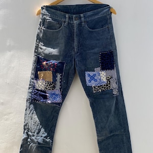 May include: Dark blue corduroy trousers with patchwork details. The patches feature various patterns, including paisley, leopard print, and floral designs. The trousers are hanging on a wooden hanger against a white background.