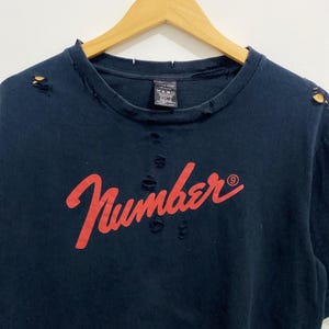 May include: A dark navy blue short-sleeve t-shirt featuring the red cursive text "Number (N)ine" across the chest. The garment has a distressed aesthetic with small holes and frayed edges along the crew neckline and shoulder seams.