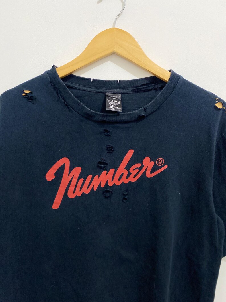 May include: A dark navy blue short-sleeve t-shirt featuring the red cursive text "Number (N)ine" across the chest. The garment has a distressed aesthetic with small holes and frayed edges along the crew neckline and shoulder seams.
