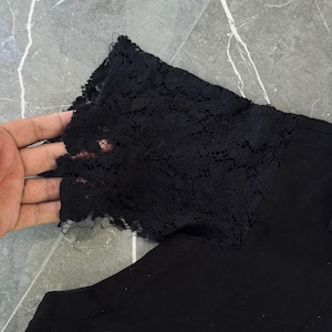 May include: Black garment with a black lace sleeve detail. The lace has a floral pattern and is attached to the sleeve. The garment is made of a solid black fabric.
