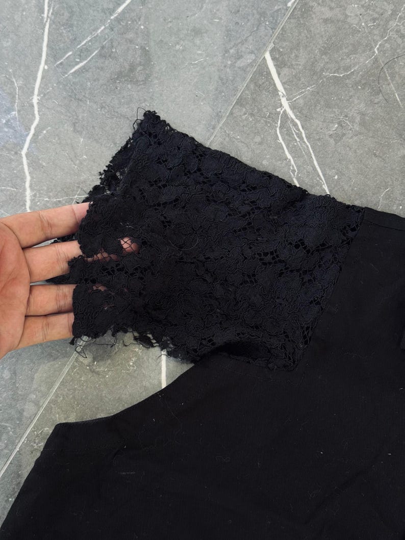 May include: Black garment with a black lace sleeve detail. The lace has a floral pattern and is attached to the sleeve. The garment is made of a solid black fabric.