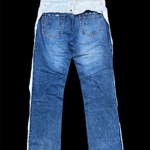 Pode incluir: Jeans azuis com cós em ganga azul clara e riscas laterais brancas. Os jeans têm dois bolsos traseiros e um design clássico de cinco bolsos. Os jeans são exibidos em um fundo preto.