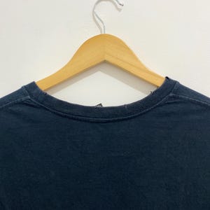 May include: A plain dark navy blue t-shirt with a crew neckline, displayed on a light-colored wooden clothes hanger. The garment features a simple, solid-colored fabric design with visible stitching along the collar and shoulder seams.