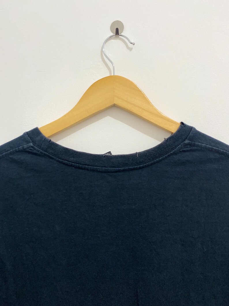 May include: A plain dark navy blue t-shirt with a crew neckline, displayed on a light-colored wooden clothes hanger. The garment features a simple, solid-colored fabric design with visible stitching along the collar and shoulder seams.