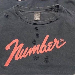 May include: A black crew neck t-shirt featuring the word Number in a red script font with a small circled nine symbol. The garment has a distressed aesthetic with multiple small holes and frayed edges around the collar and across the chest area.