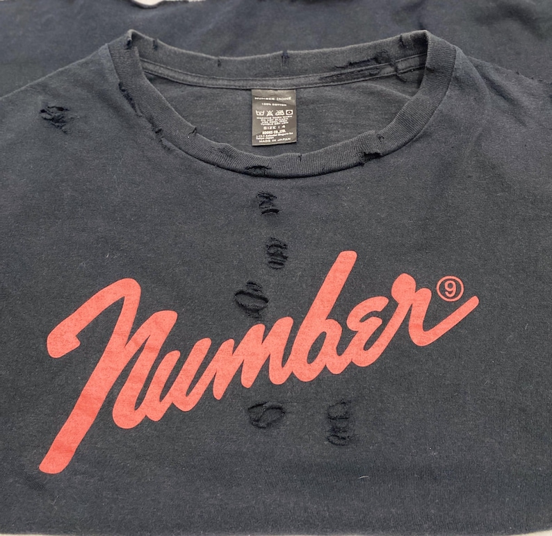 May include: A black crew neck t-shirt featuring the word Number in a red script font with a small circled nine symbol. The garment has a distressed aesthetic with multiple small holes and frayed edges around the collar and across the chest area.