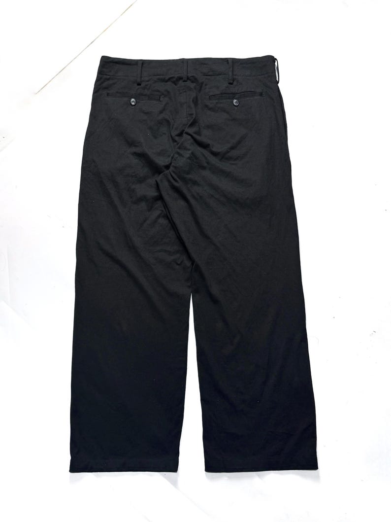 Puede incluir: Pantalones negros de pernera ancha con dos bolsillos traseros y cierres de bot&oacute;n. Los pantalones tienen un dise&ntilde;o cl&aacute;sico y est&aacute;n hechos de una tela ligera. Los pantalones se muestran desde la parte trasera sobre un fondo blanco.