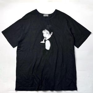 May include: Black short-sleeved t-shirt featuring a black and white illustration of a person with short hair, with a finger to their lips. The graphic is centered on the front of the shirt. The shirt has a crew neck.