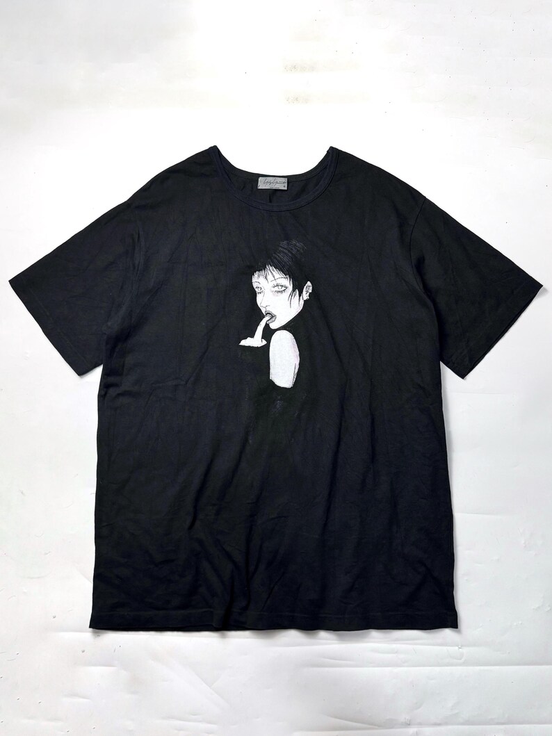 May include: Black short-sleeved t-shirt featuring a black and white illustration of a person with short hair, with a finger to their lips. The graphic is centered on the front of the shirt. The shirt has a crew neck.