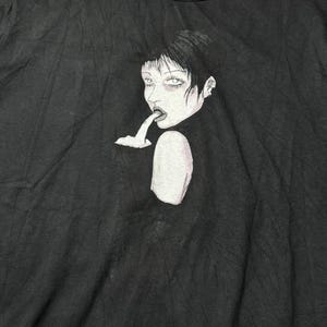 May include: Black crew neck t-shirt featuring a graphic illustration of a woman with dark hair and pale skin. The artwork depicts a woman with her finger in her mouth. The shirt has a small tag at the neckline.