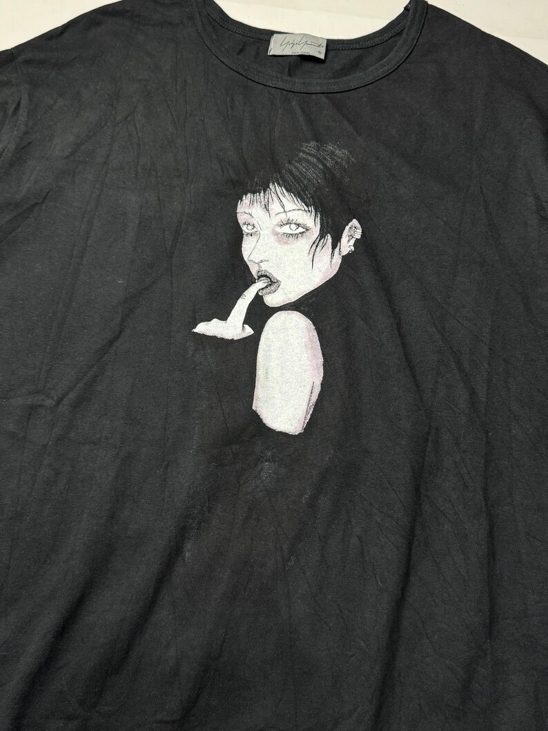 May include: Black crew neck t-shirt featuring a graphic illustration of a woman with dark hair and pale skin. The artwork depicts a woman with her finger in her mouth. The shirt has a small tag at the neckline.