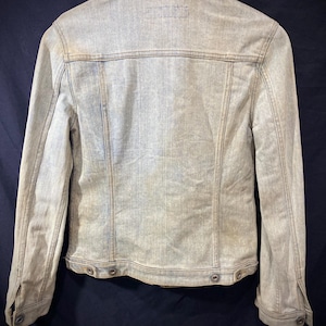 May include: A light-wash denim jacket shown from the back, featuring a classic collar, long sleeves with buttoned cuffs, vertical seam detailing, and a bottom hem with adjustable button tabs on each side, presented on a wooden hanger.