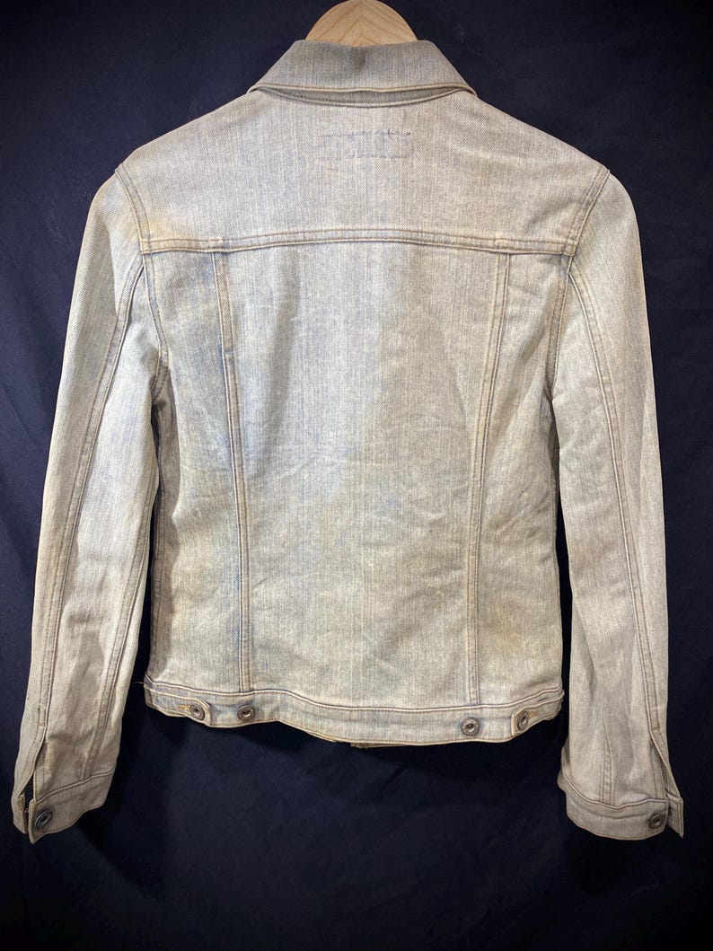 May include: A light-wash denim jacket shown from the back, featuring a classic collar, long sleeves with buttoned cuffs, vertical seam detailing, and a bottom hem with adjustable button tabs on each side, presented on a wooden hanger.