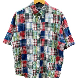 May include: Short-sleeved patchwork shirt in a variety of plaid patterns, including red, blue, green, and white. The shirt has a button-down collar and a small embroidered logo on the chest. The shirt is hanging on a wooden hanger.