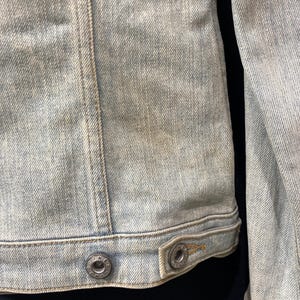 May include: A light blue denim jacket featuring a faded wash, vertical seam detailing, and a bottom hem with two metallic buttons. Each button is embossed with the text "THE DENIM" around the center, providing a secure closure for the garment.