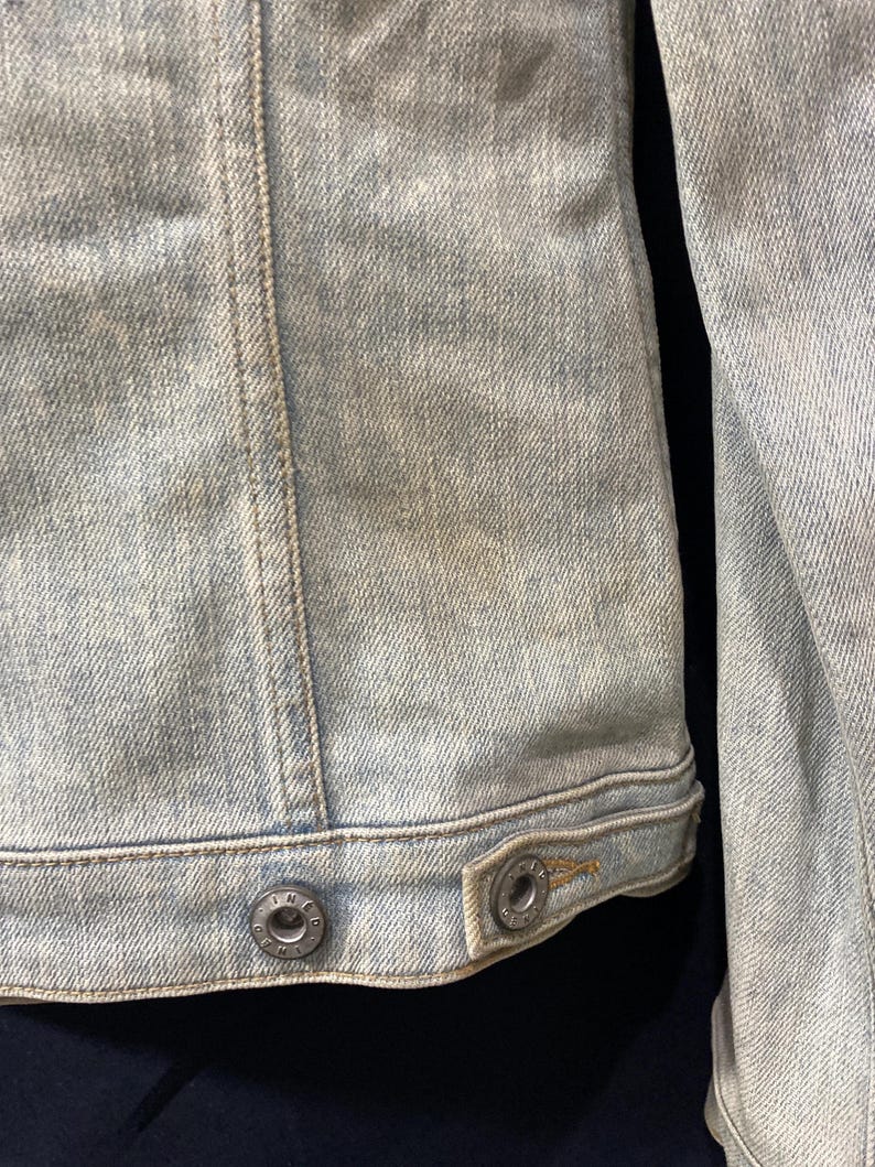 May include: A light blue denim jacket featuring a faded wash, vertical seam detailing, and a bottom hem with two metallic buttons. Each button is embossed with the text "THE DENIM" around the center, providing a secure closure for the garment.