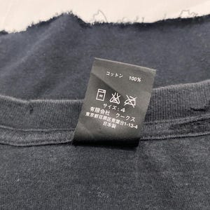 May include: A black garment care label featuring white text and laundry symbols. The label indicates 100% cotton, size 4, and includes Japanese text detailing the manufacturer, Coo Coos Ltd., located in Meguro-ku, Tokyo, and states it is made in Japan.