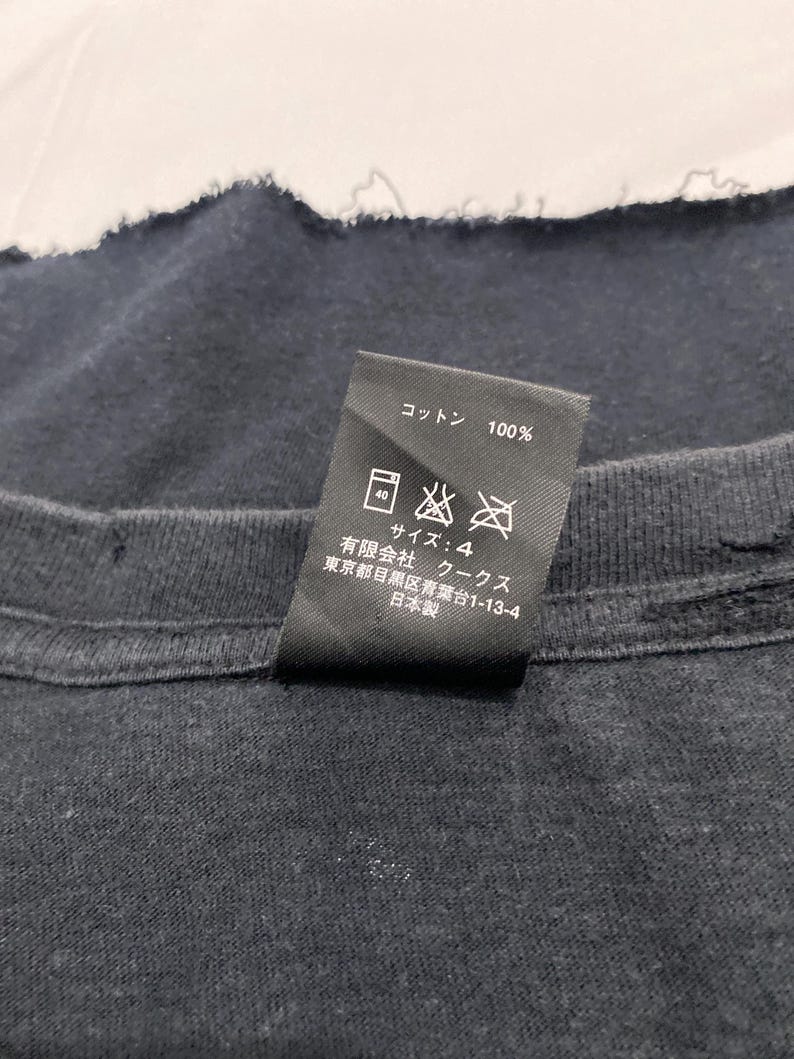 May include: A black garment care label featuring white text and laundry symbols. The label indicates 100% cotton, size 4, and includes Japanese text detailing the manufacturer, Coo Coos Ltd., located in Meguro-ku, Tokyo, and states it is made in Japan.