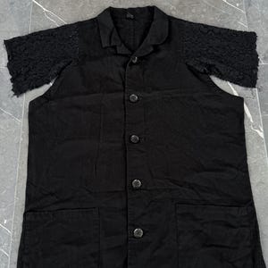 May include: Black button-down shirt with short sleeves. The sleeves are made of a textured, open-weave lace. The shirt has a collar, two front pockets, and four buttons down the front. The fabric appears to be a medium-weight cotton or linen.