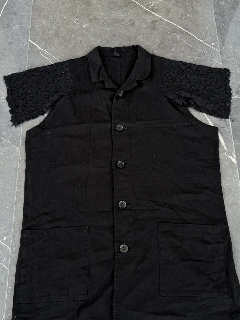May include: Black button-down shirt with short sleeves. The sleeves are made of a textured, open-weave lace. The shirt has a collar, two front pockets, and four buttons down the front. The fabric appears to be a medium-weight cotton or linen.
