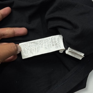 May include: Close-up of a black t-shirt with a tag attached. The tag contains text and care symbols. The shirt is made of a soft material. The image is well-lit, showing the details of the fabric and the tag.