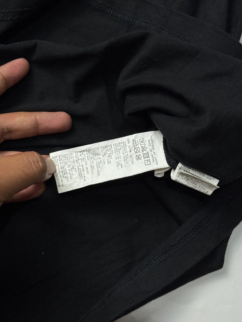 May include: Close-up of a black t-shirt with a tag attached. The tag contains text and care symbols. The shirt is made of a soft material. The image is well-lit, showing the details of the fabric and the tag.