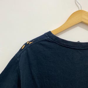 May include: A dark navy blue t-shirt featuring a crew neckline and short sleeves. The shoulder seam on the left side displays visible wear with small, irregular holes along the fabric edge, showing signs of fraying and material distress.