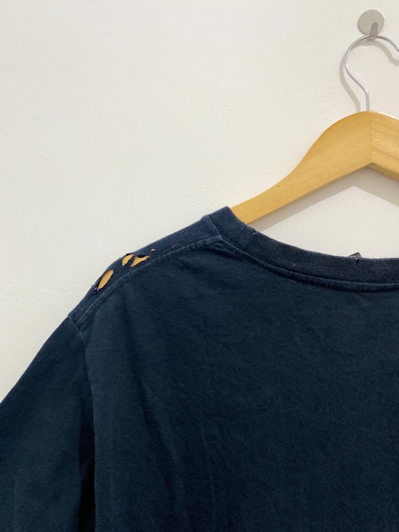May include: A dark navy blue t-shirt featuring a crew neckline and short sleeves. The shoulder seam on the left side displays visible wear with small, irregular holes along the fabric edge, showing signs of fraying and material distress.