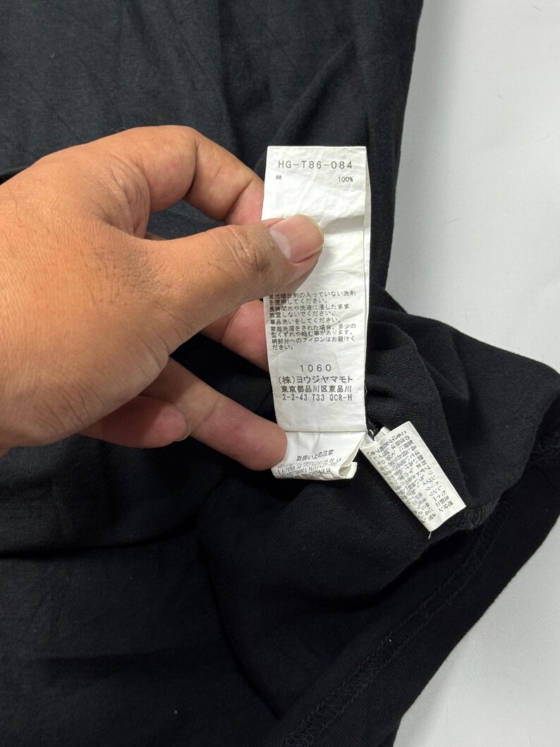 May include: Black t-shirt with a white tag. The tag has the text "HG-T86-084" and "100%" printed on it. The tag also has Japanese text and the number "1060".
