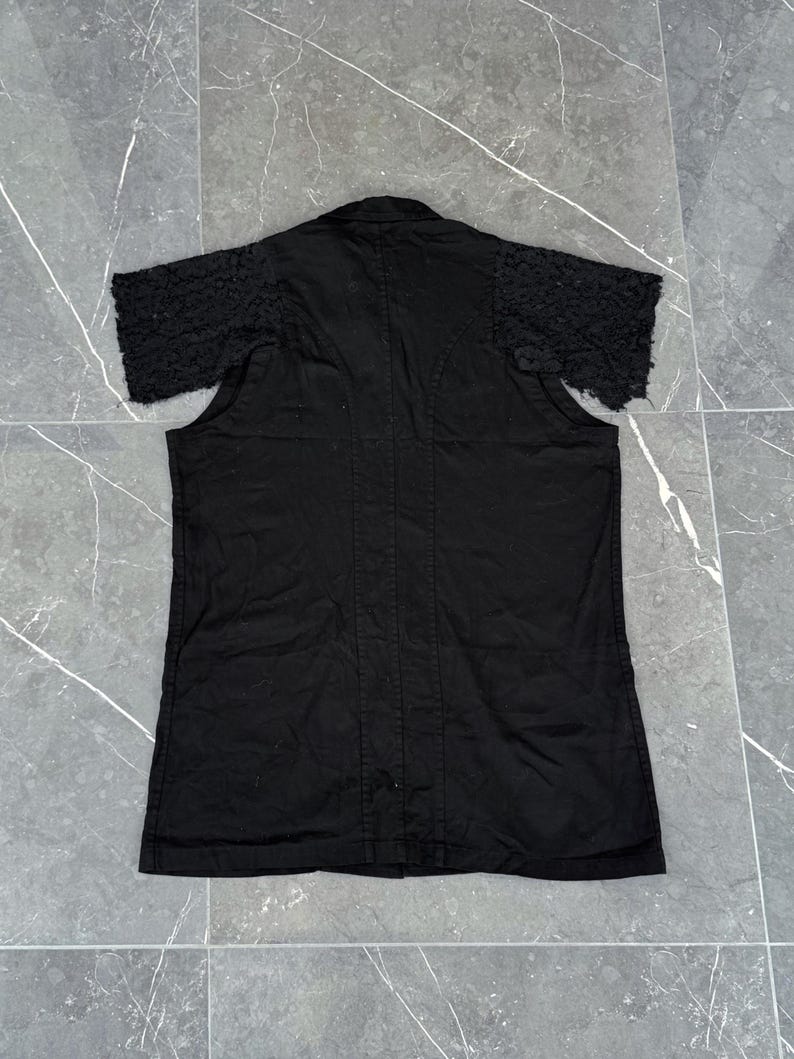 May include: Black short-sleeved shirt with textured sleeves. The shirt has a button-down front and a collar. The sleeves have a unique, textured design. The shirt is made of a dark fabric.