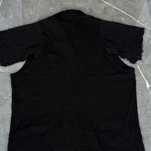 May include: Black short-sleeved jacket with textured sleeves. The jacket has a collar and a vertical seam down the center of the back. The sleeves have a unique, textured design, adding visual interest to the garment. The jacket is made of a solid black fabric.