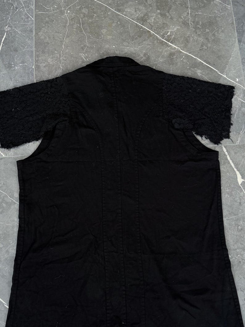 May include: Black short-sleeved jacket with textured sleeves. The jacket has a collar and a vertical seam down the center of the back. The sleeves have a unique, textured design, adding visual interest to the garment. The jacket is made of a solid black fabric.