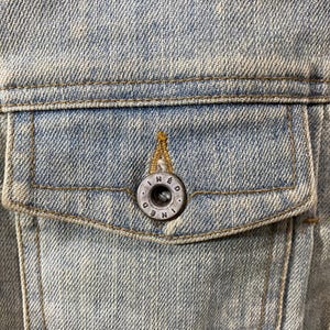 May include: A light blue denim jacket pocket featuring a weathered texture and gold-toned stitching. A circular, dark metallic button is centered on the pocket flap, embossed with the text INED in a circular pattern around the center hole.